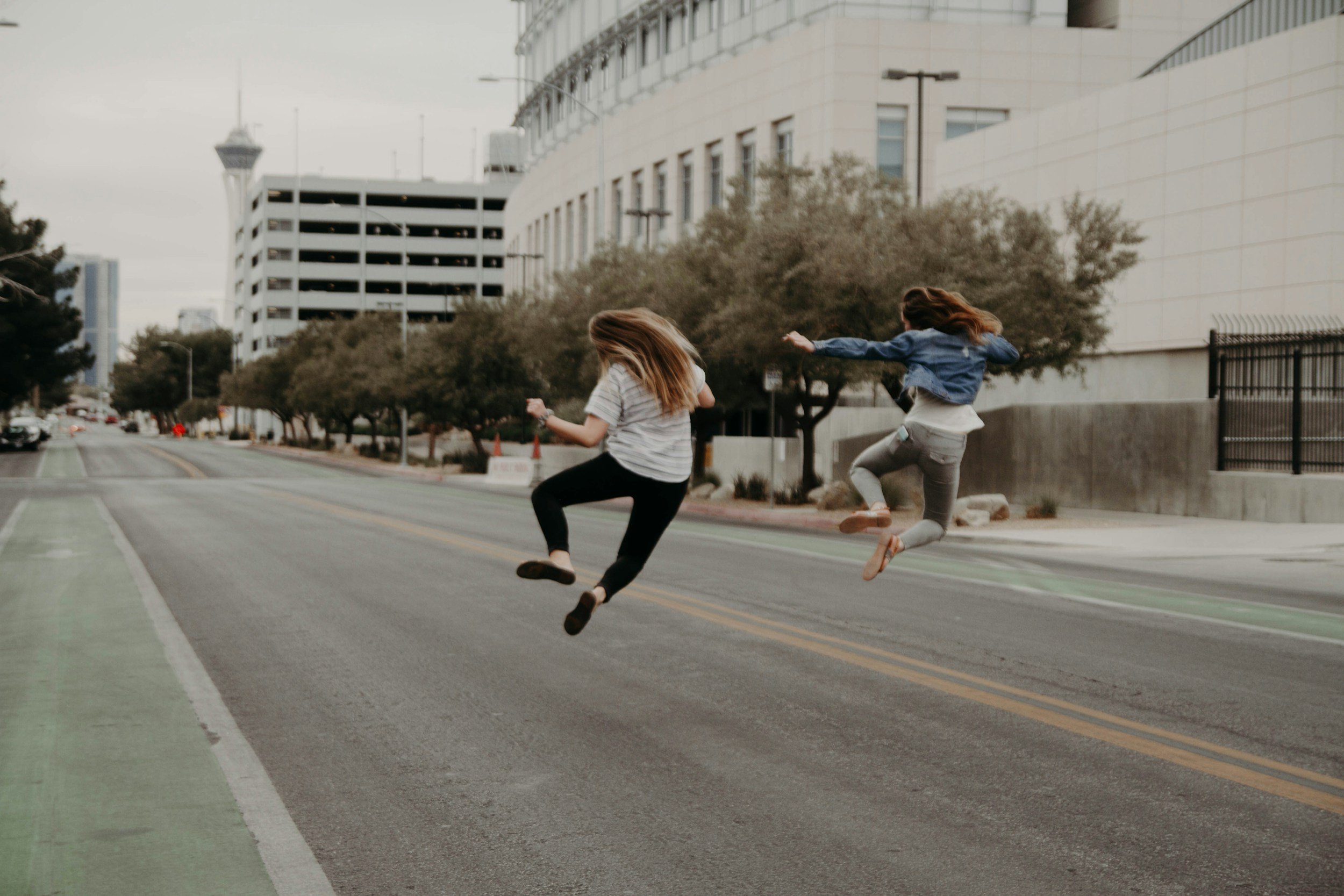 Two girls jumping on an empty city street with modern buildings and trees in the background.
