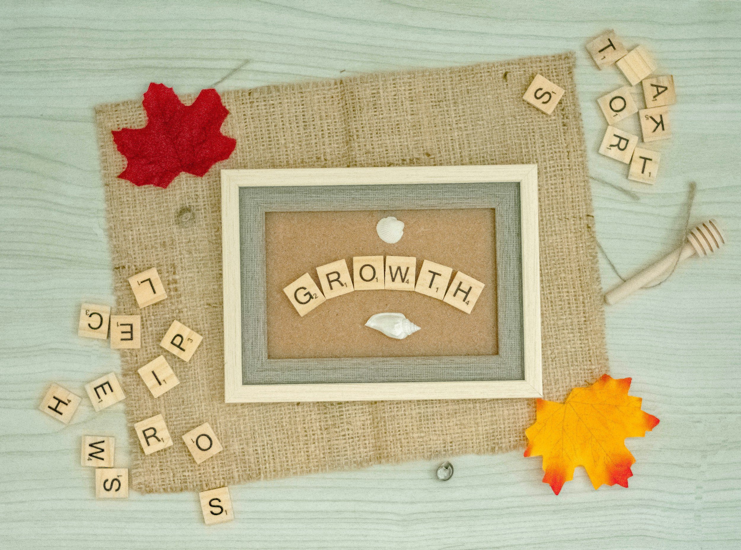 A framed display with the words "GROWTH" on letter tiles, surrounded by artificial fall leaves, scattered letter tiles, a small bean uses for a loom, and a piece of burlap fabric on a wooden surface.